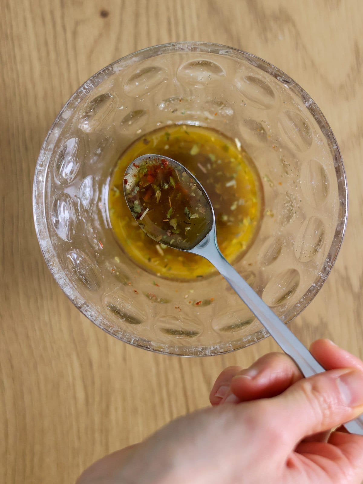 A hand holds a teaspoon with olive oil dressing on it. Under the spoon is a bowl with more dressing.