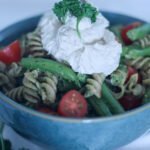 Pasta with green pesto, cherry tomatoes, green beans and vegan ricotta cheese in a blue bowl.