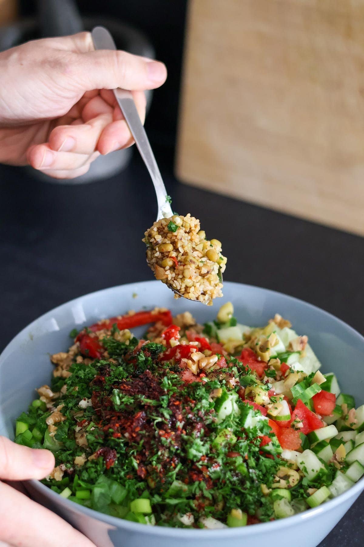 A hand holds a spoon with bulgur and mung bean salad above a blue bowl filled with more salad.