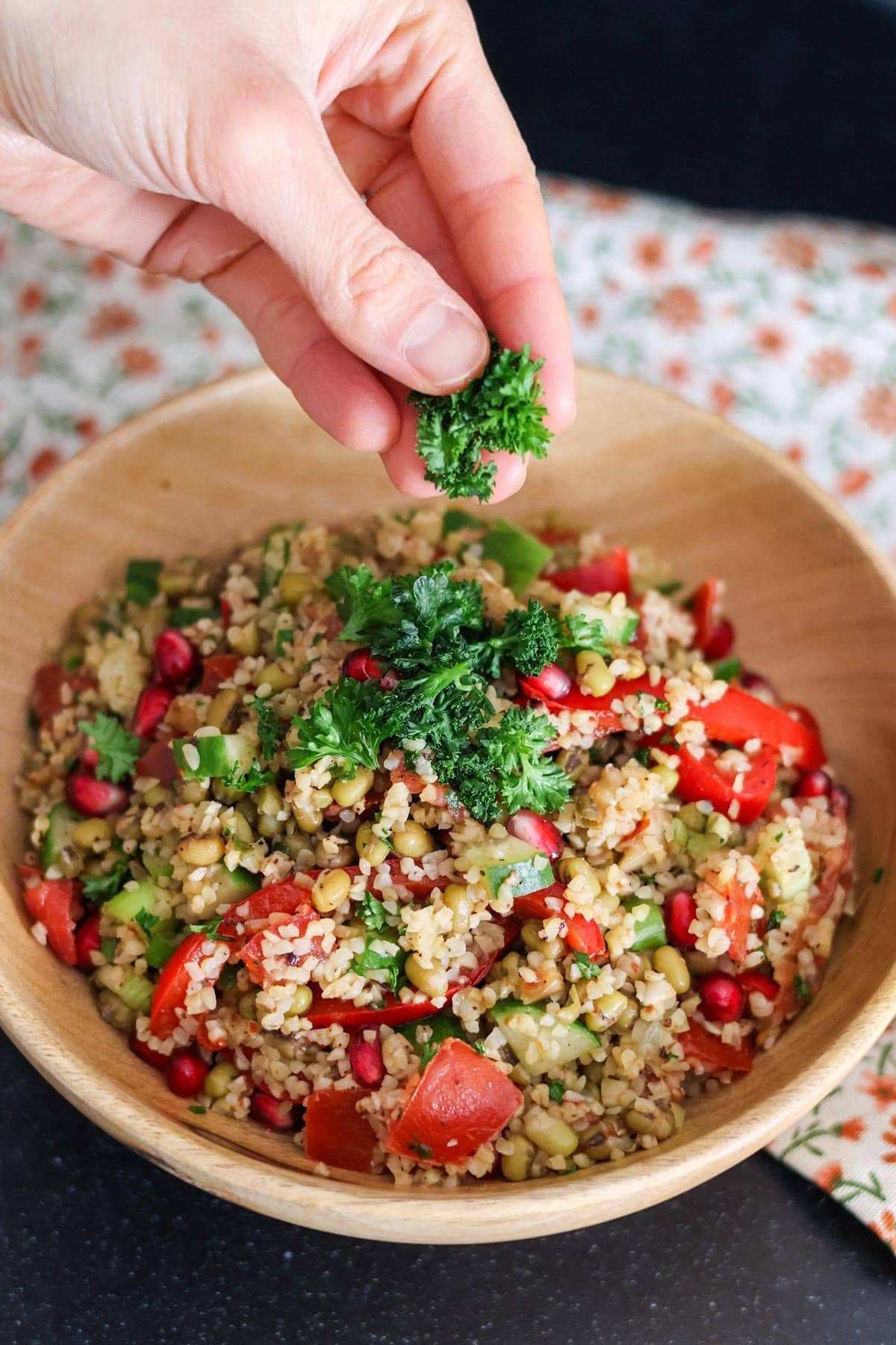 a hand puts fresh parsley on the salad in a wooden bowl.