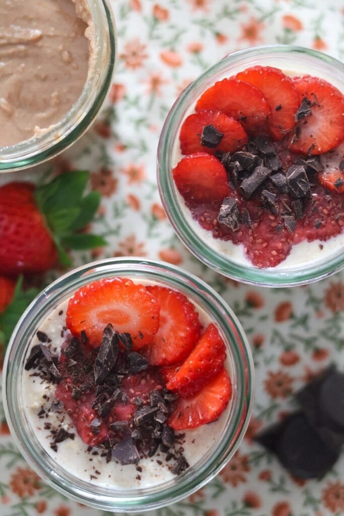 Overnight oats in two glass jars topped with strawberry jelly, fresh strawberries and chocolate.