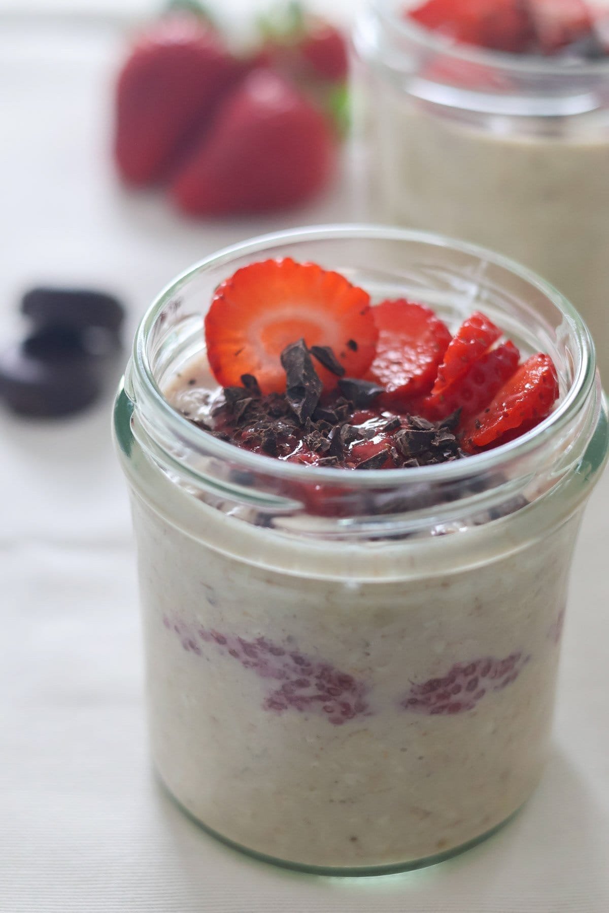 A jar of glass filled with pb and jelly overnight oats with dark chocolate and fresh strawberries in the back.