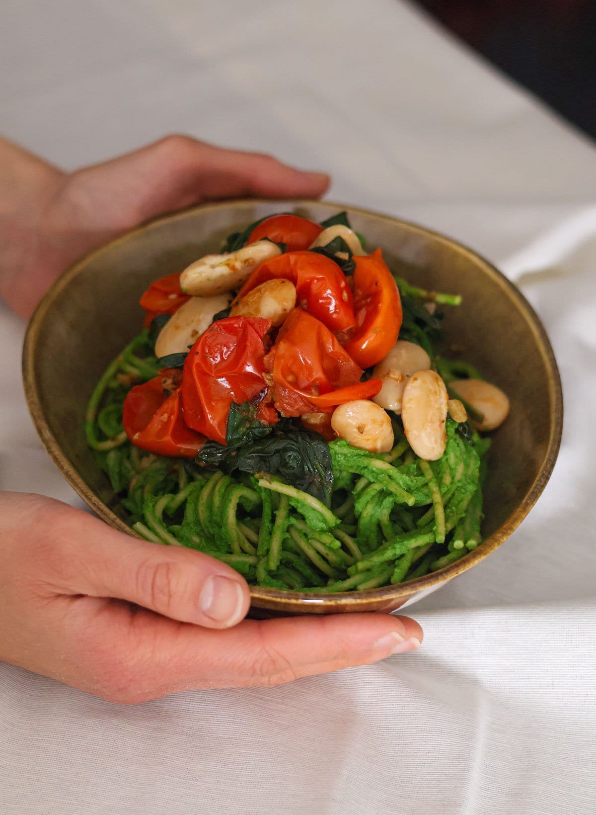 Two hands hold a brown bowl with spaghetti with a green sauce.