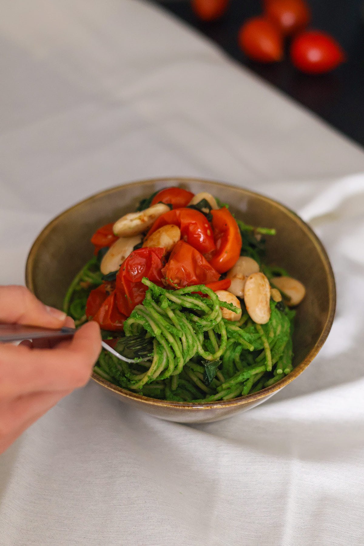 A fork in a brown bowl filled with green spaghetti topped with tomatoes and butter beans. 