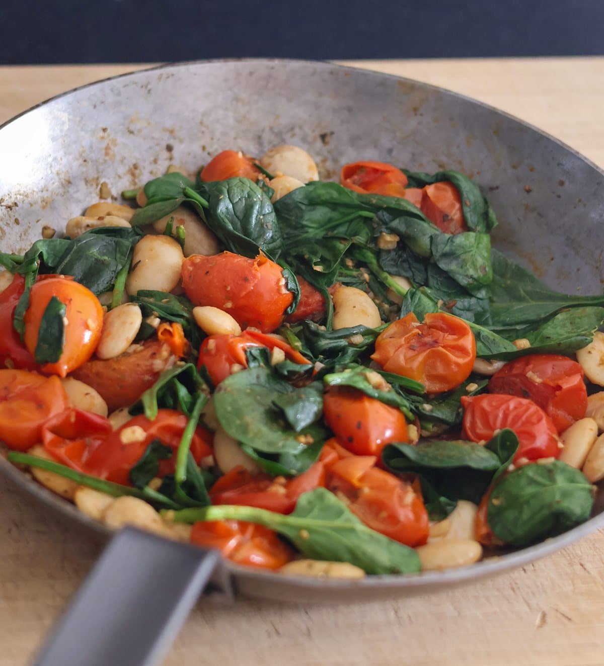 A pan on a wooden board with fried tomatoes, butter beans and spinach.