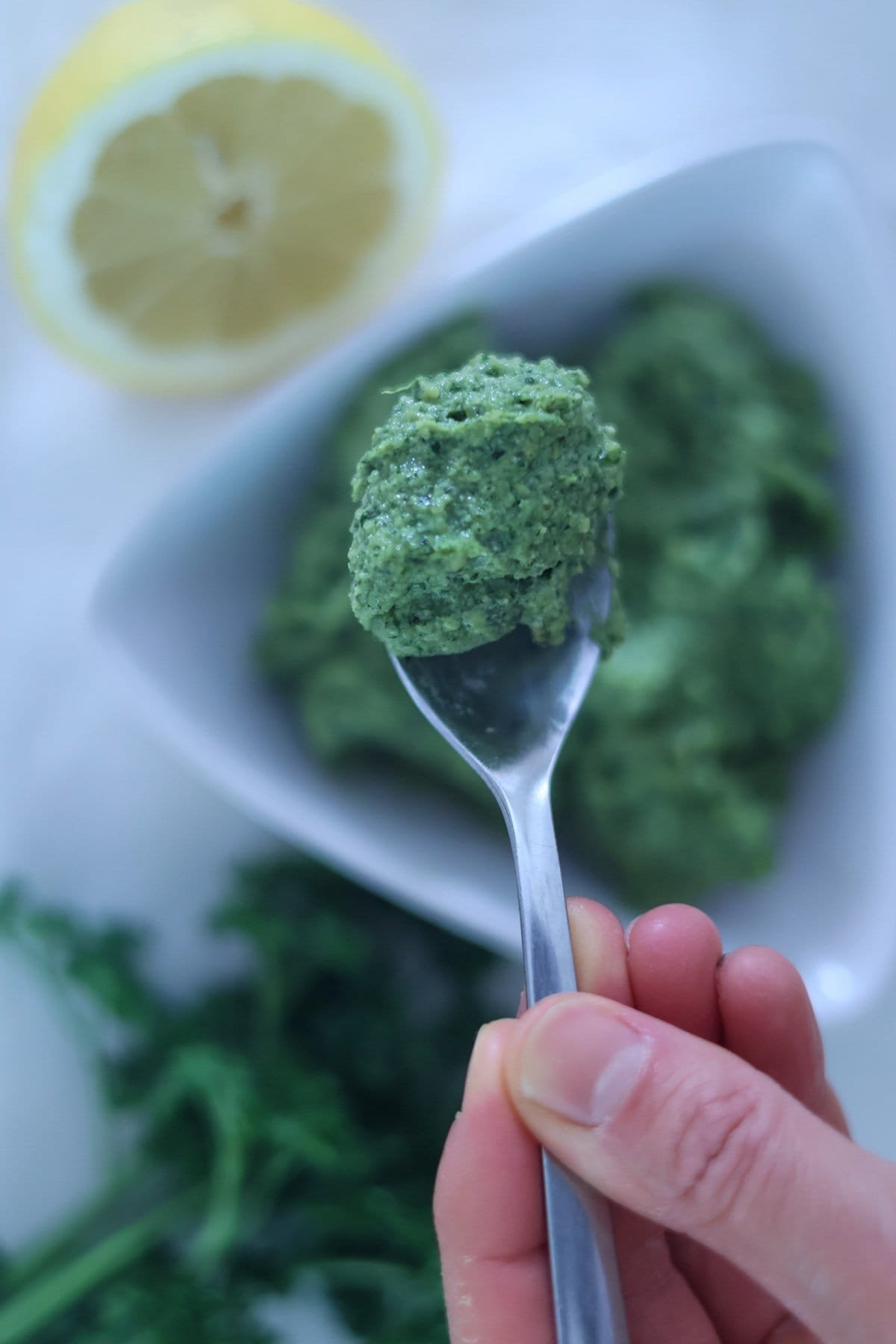 A hand holds a silver spoon with vegan green pesto.