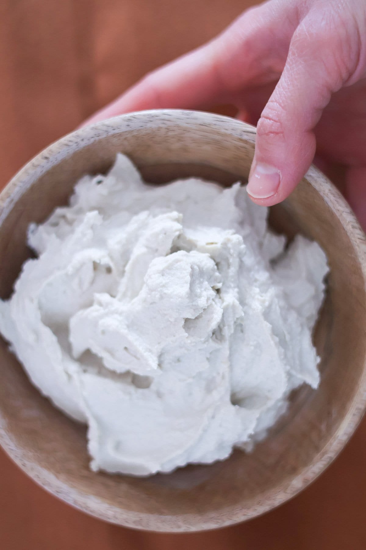 A hand holds a wooden bowl filled with vegan ricotta cheese.