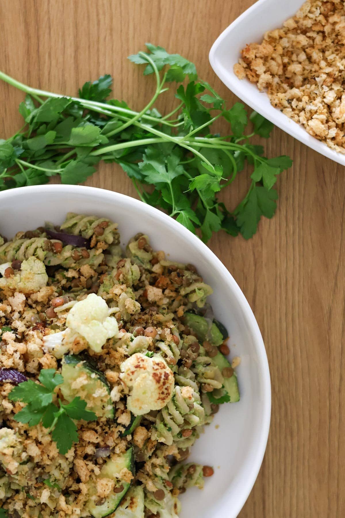 Vegan pasta salad in a white bowl on a wooden table with fresh parsley and breadcrumbs on the side.