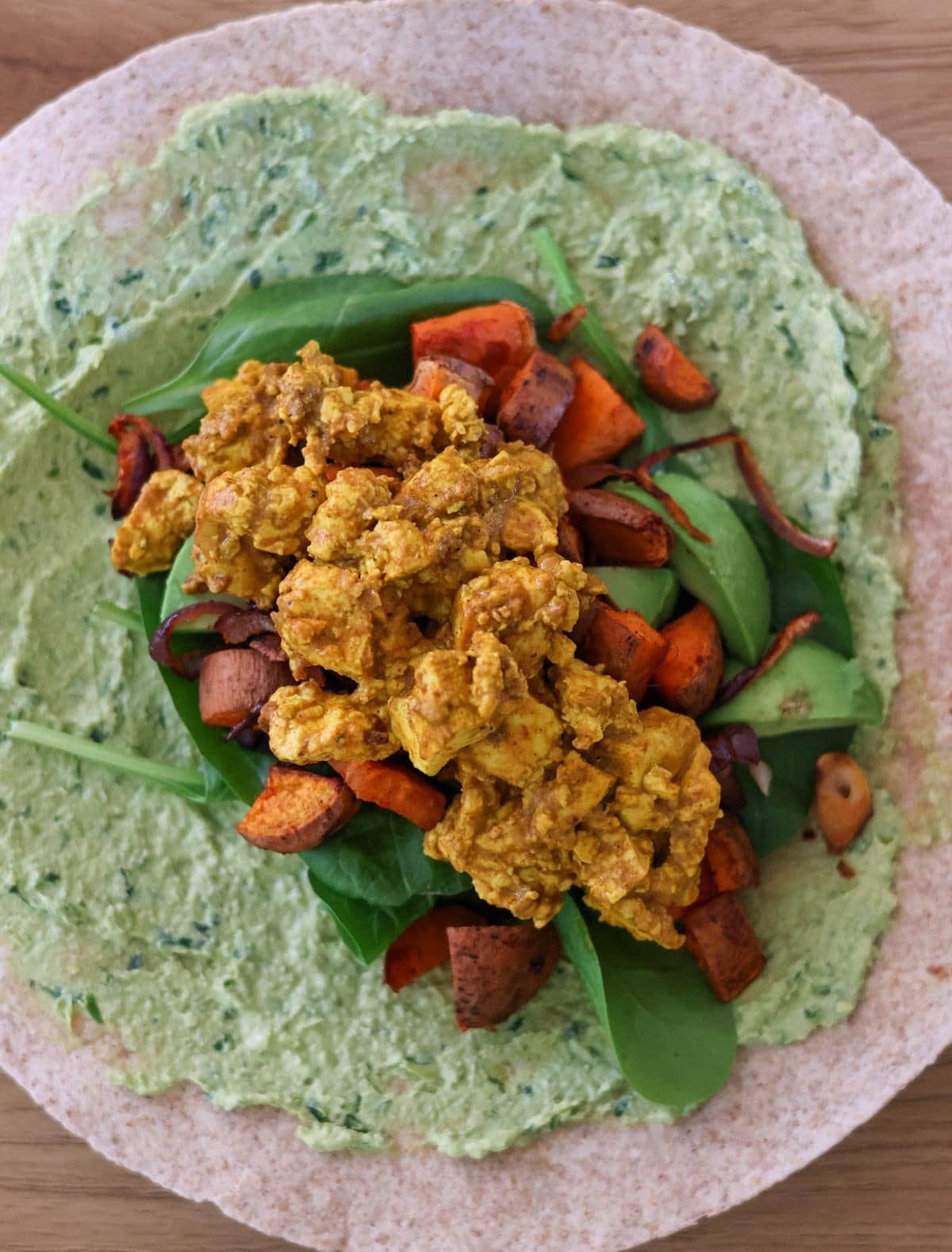 a wrap topped with scrambled tofu, sweet potato, red onion, spinach, avocado and a green sauce.