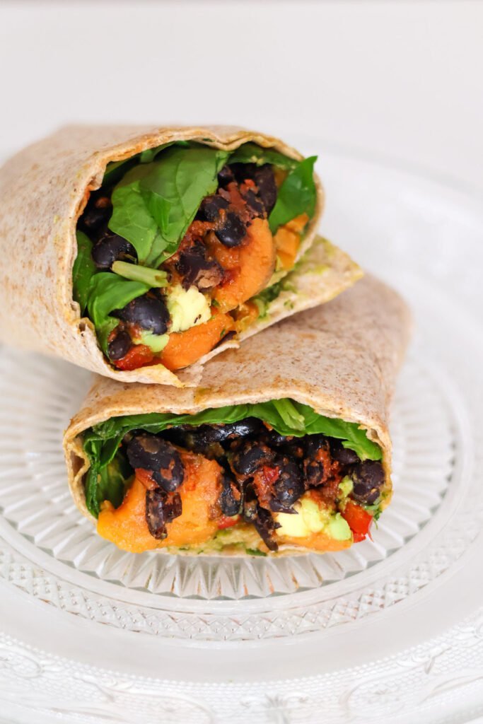 A wrap cut in half filled with black beans, sweet potato and spinach lies on a plate.