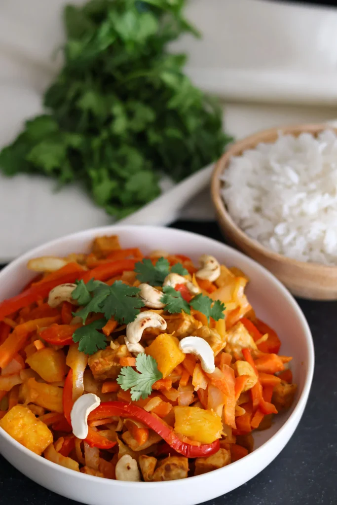 Pineapple and cashew stir-fry in a white bowl with a bowl of rice and a bunch of fresh cilantro in the background.