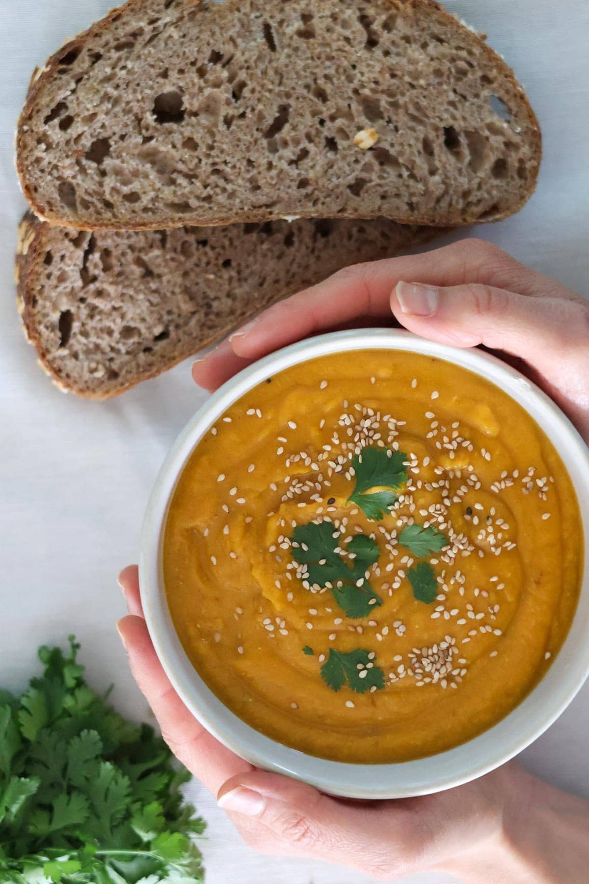 Two hands hold a white bowl filled with carrot sweet potato and ginger soup topped with cilantro and sesame seeds.
