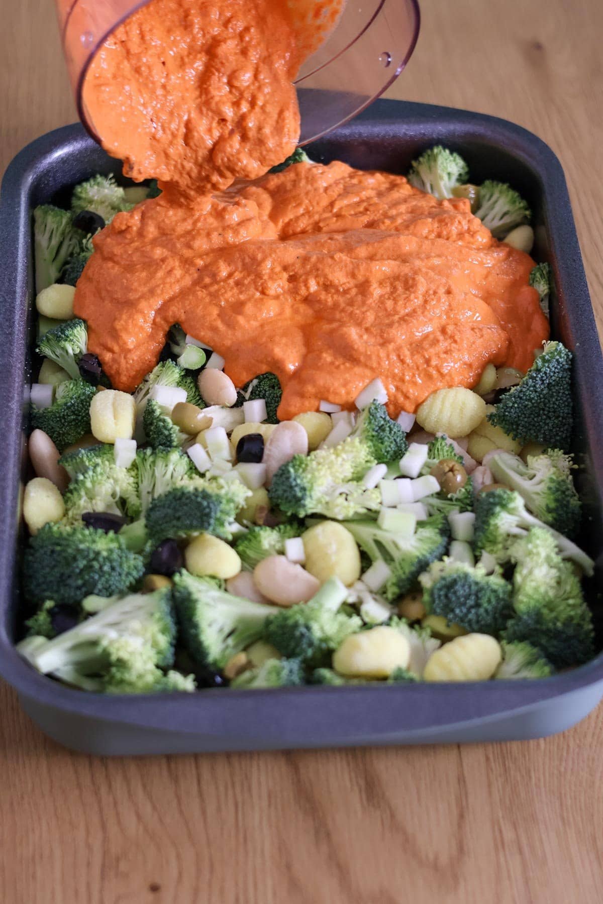 An orange sauce is poured over a baking dish of uncooked gnocchi, broccoli, butter beans and olives.