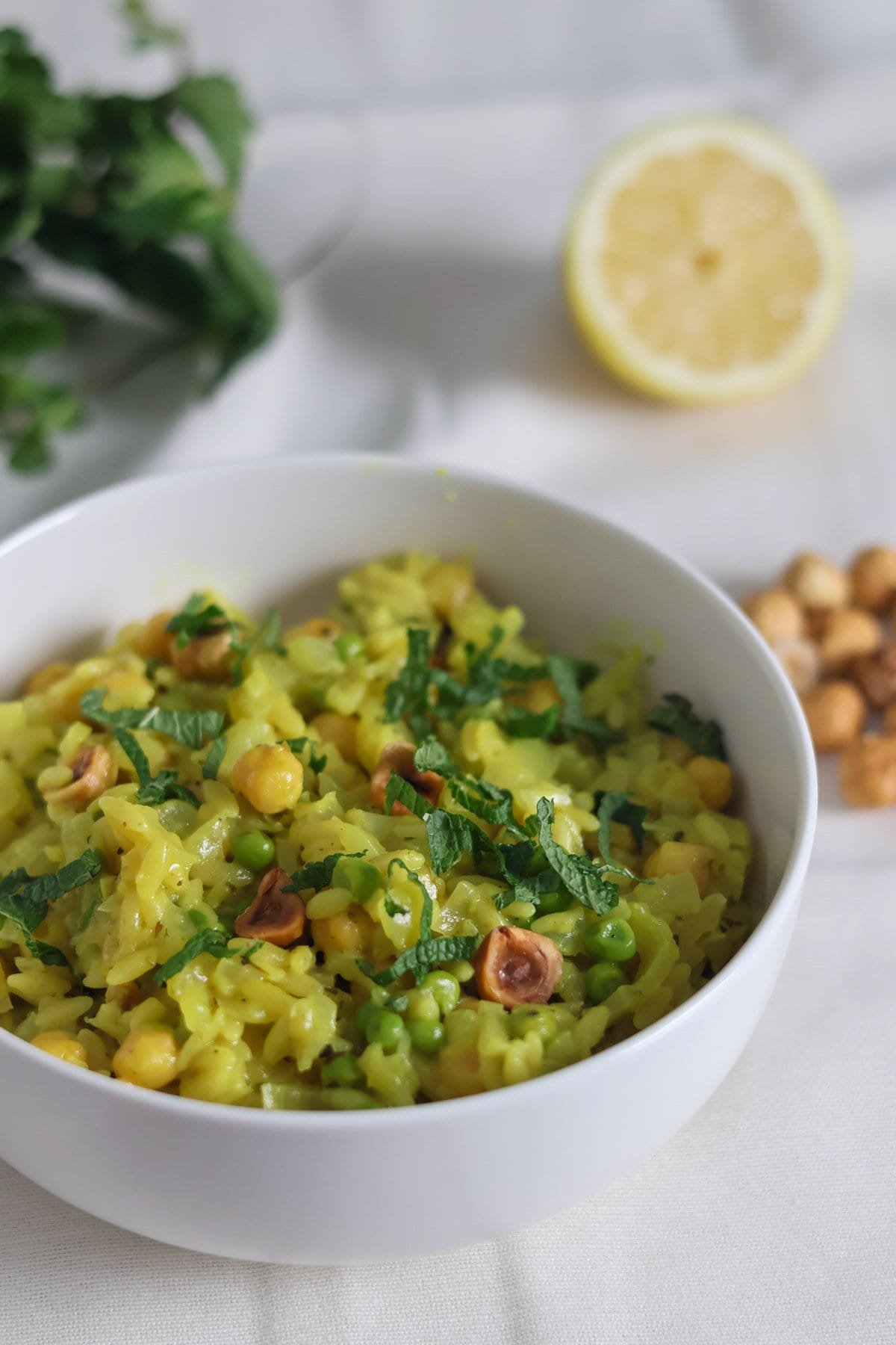 Orzo with chickpeas, lemon, fresh mint and hazelnuts in a white bowl with more hazelnuts, fresh mint and lemon in de background.