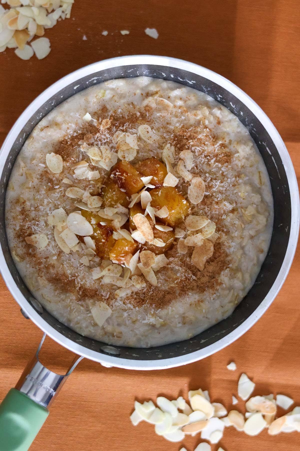 Oatmeal in a green saucepan topped with roasted pineapple, cinnamon, shredded coconut and slivered toasted almonds.