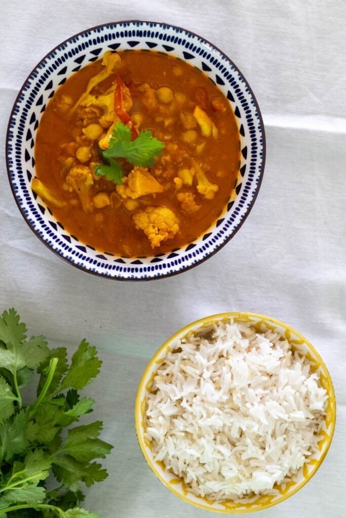 Cauliflower and chickpea curry in a blue and white bowl served with fresh cilantro and rice in another bowl on the side.