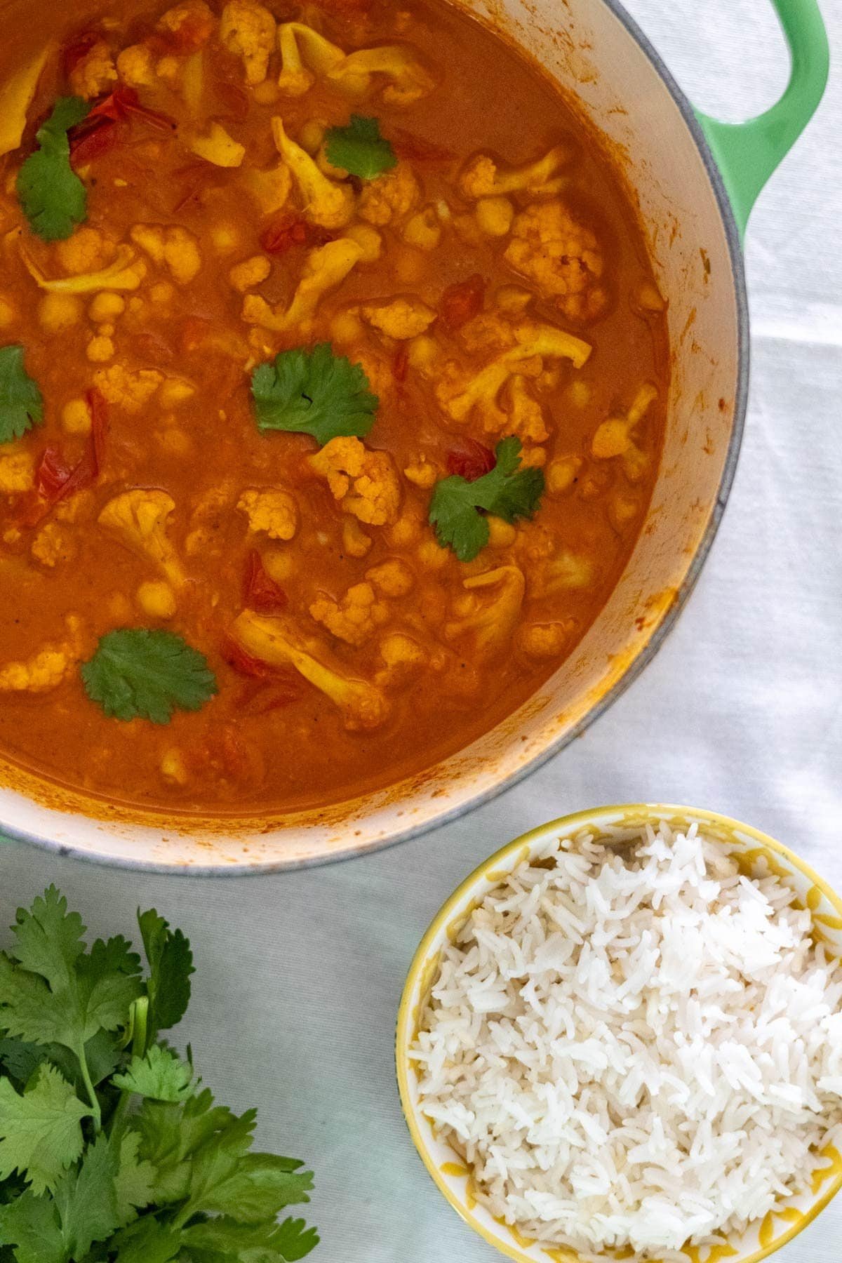 Cauliflower and chickpea curry in a large green pot with fresh cilantro and rice in a bowl on the side.