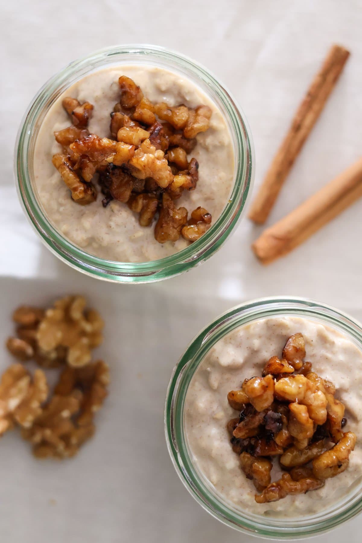 Two jars filled with cinnamon overnight oats and caramelized walnuts with walnuts and cinnamon sticks in the background.