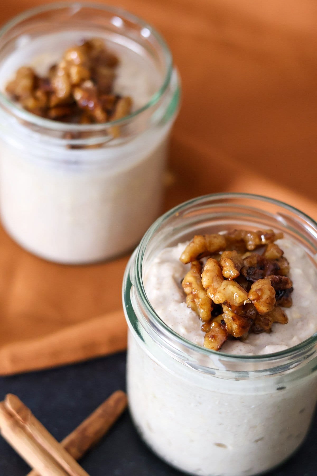 Two jars filled with cinnamon roll overnight oats and walnuts with an orange cloth in the background.