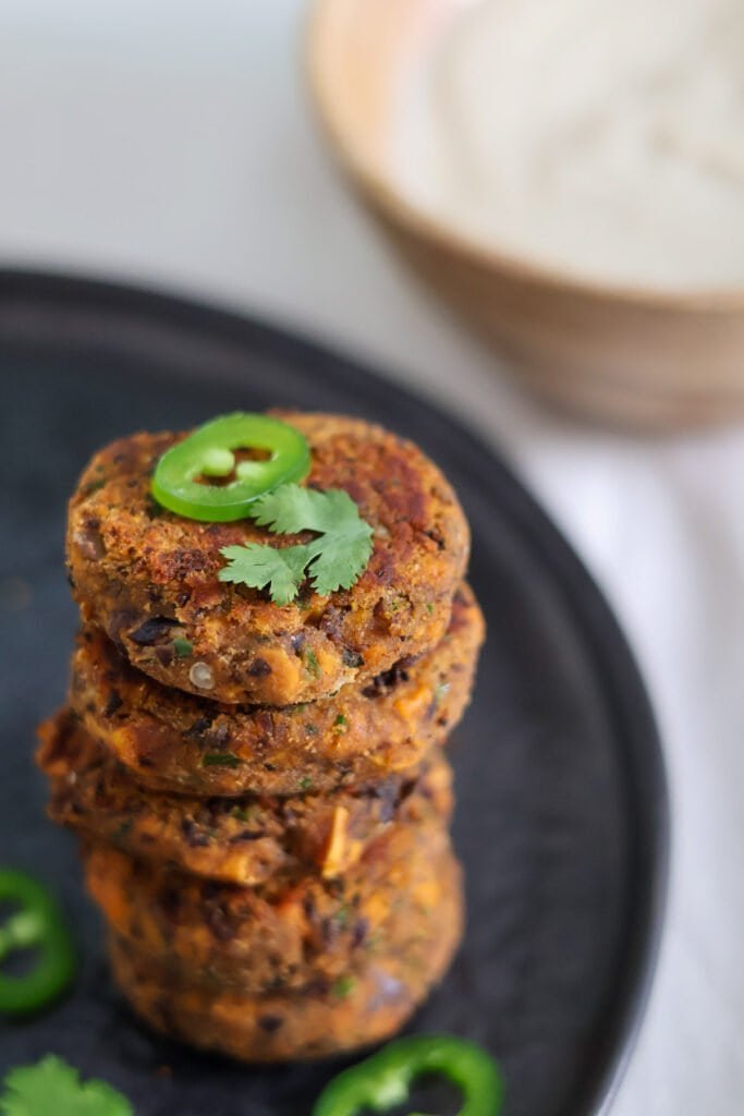Vegan sweet potato cakes stacked on top of each other on a black plate finished with fresh cilantro and jalapeño pepper.