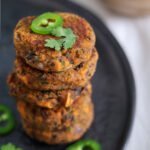 Vegan sweet potato cakes stacked on top of each other on a black plate finished with fresh cilantro and jalapeño pepper.