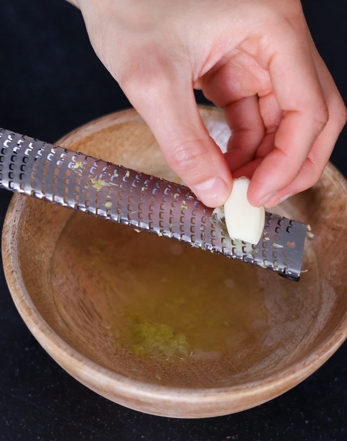 A hand grates a clove of garlic above a wooden bowl filled with lemon juice and zest.