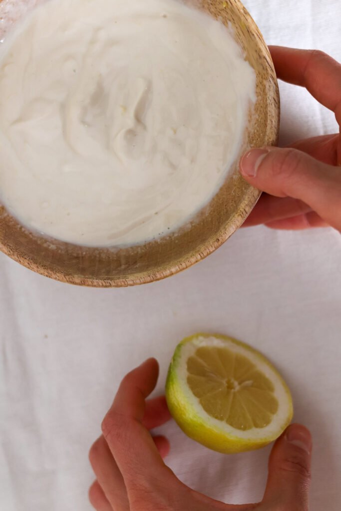 One hand holds a wooden bowl with lemon tahini yogurt sauce, another hand grabs half a lemon.
