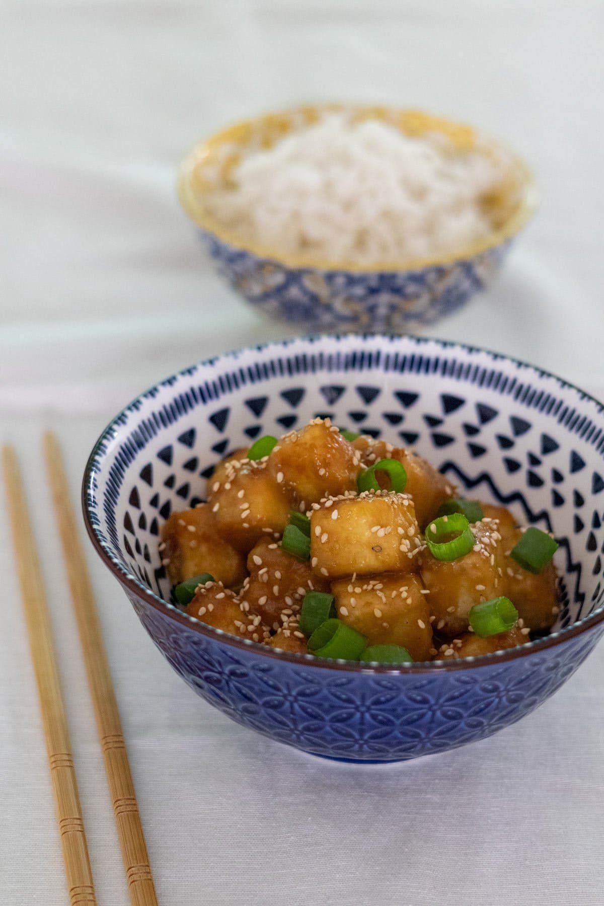 Orange tofu topped with sesame seeds and spring onions in a blue bowl with chopsticks and rice on the side.