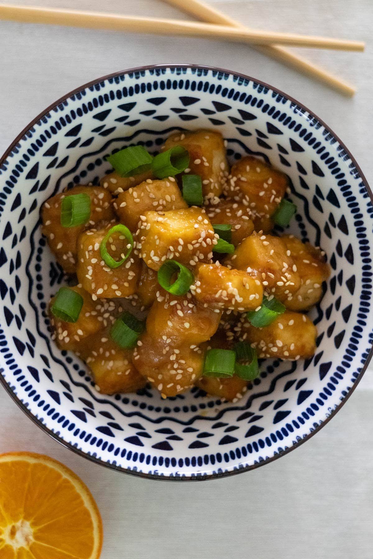 Orange tofu topped with spring onions and sesame seeds in a blue and white bowl with chopsticks and half an orange on the side.