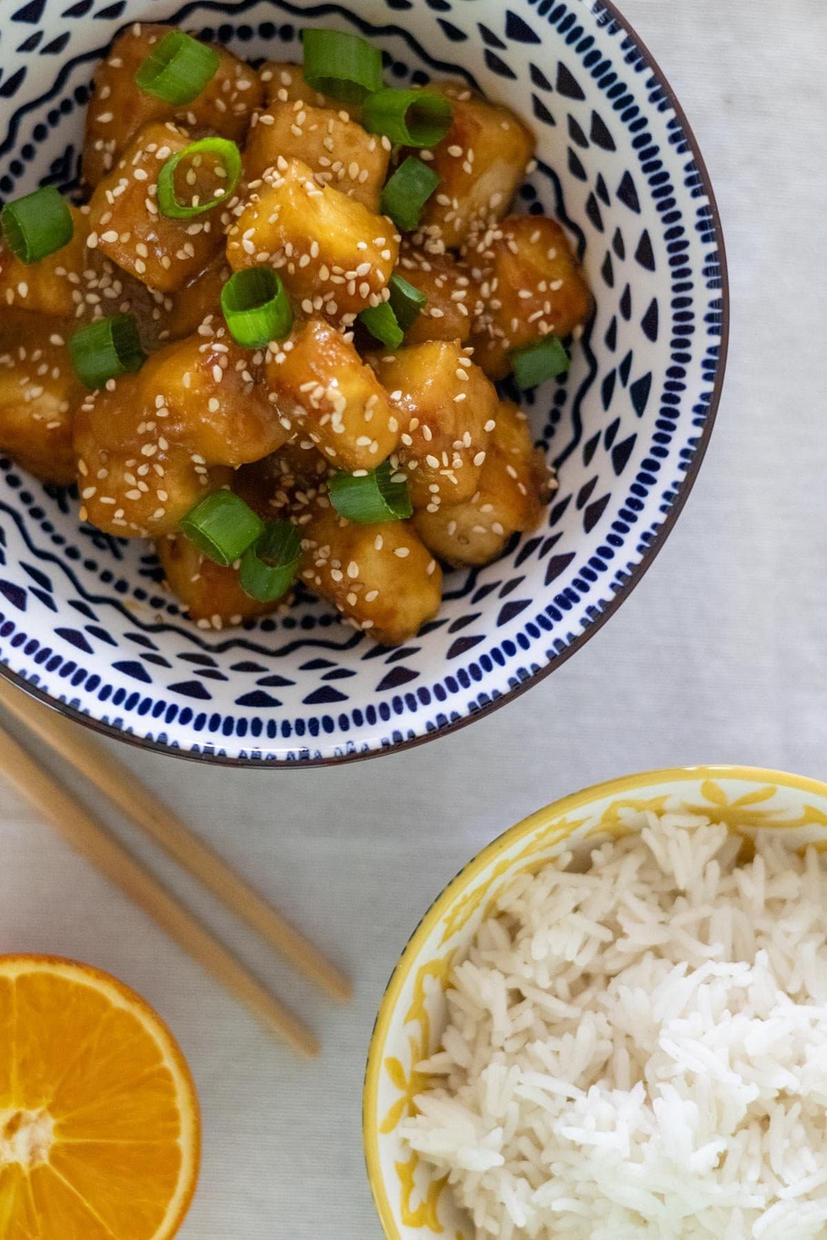 Orange tofu topped with spring onions and sesame seeds in a blue and white bowl with rice and half an orange on the side.