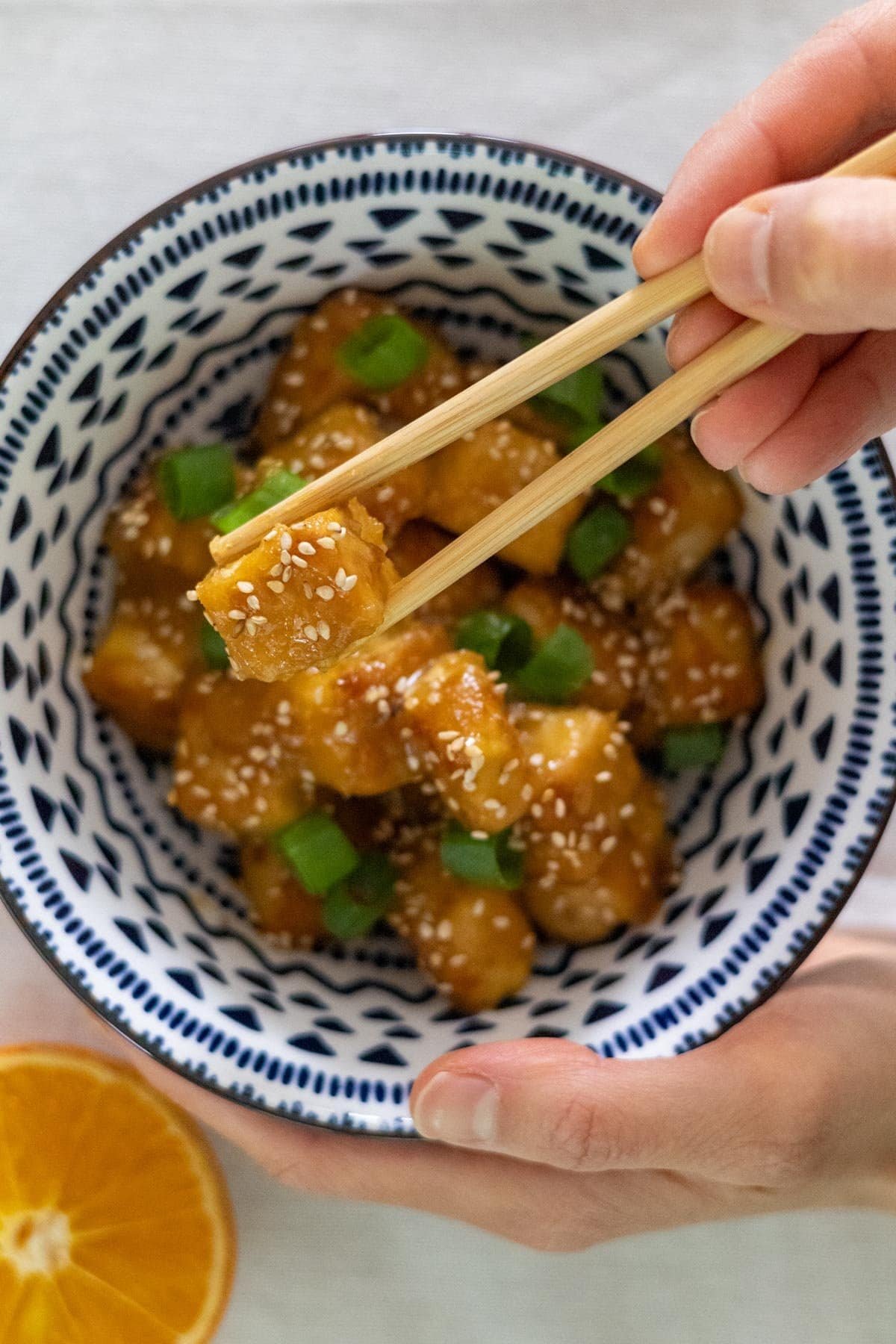 A hand picks up a piece of orange tofu with chopsticks.