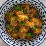 Orange tofu topped with spring onions and sesame seeds in a blue and white bowl.