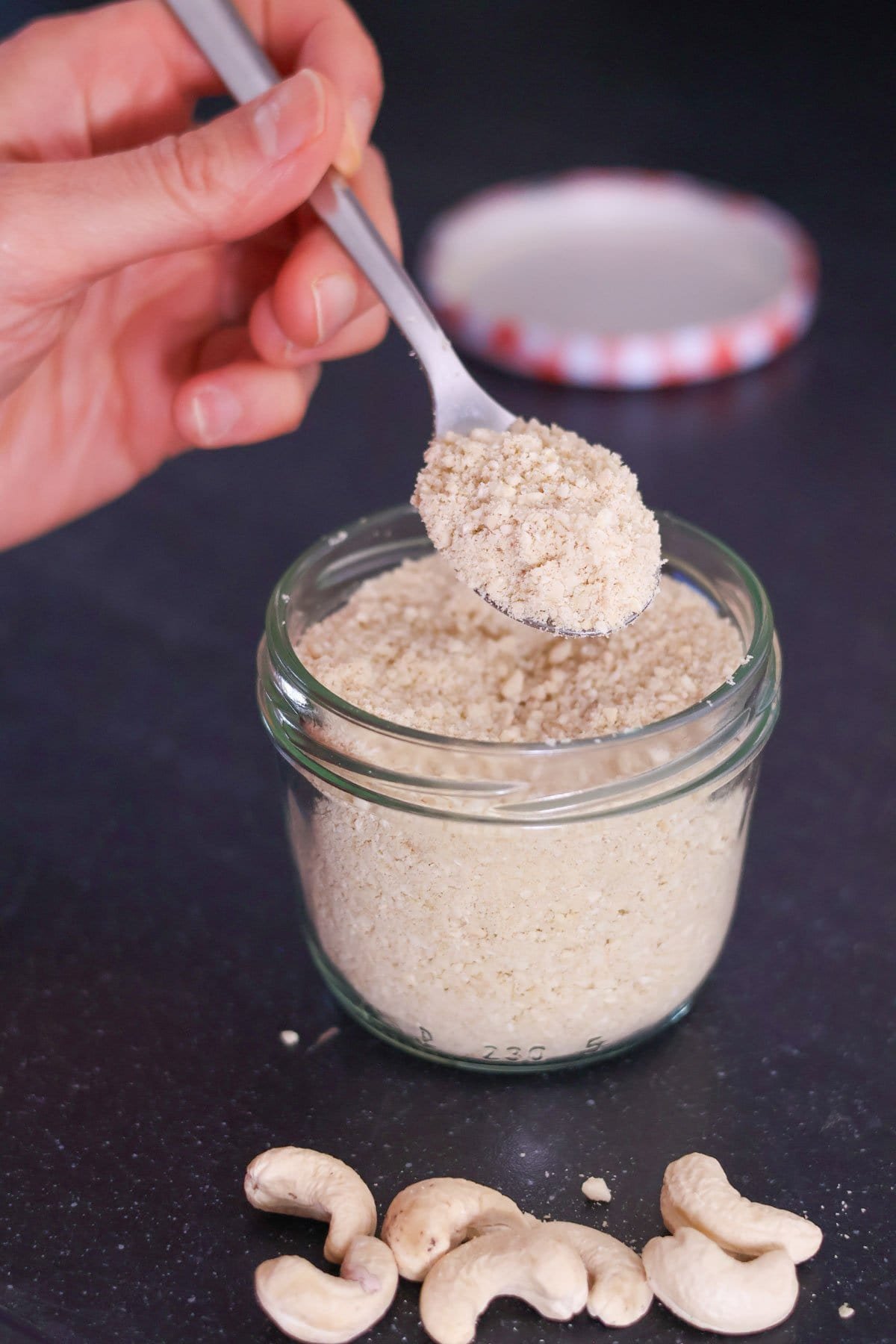 A hand holds a silver spoon with vegan parmesan cheese above a jar with more vegan parmesan cheese.