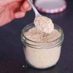 A hand holds a silver spoon with vegan parmesan cheese above a jar with more vegan parmesan cheese.
