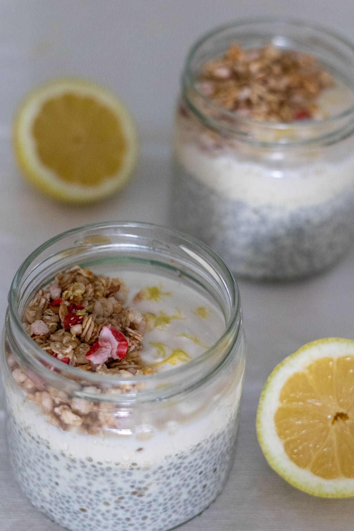 Two small mason jars filled with chia pudding topped with plant-based yogurt, granola and lemon zest with two half lemons on the side.