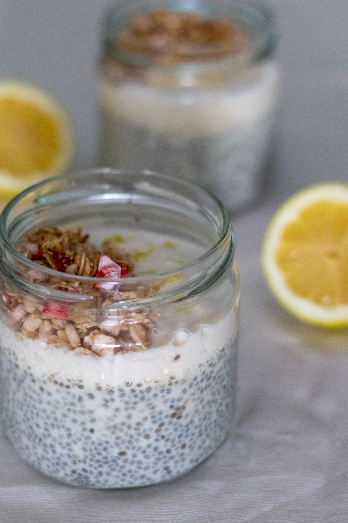 Two small mason jars filled with chia pudding topped with plant-based yogurt, granola and lemon zest with two half lemons on the side.