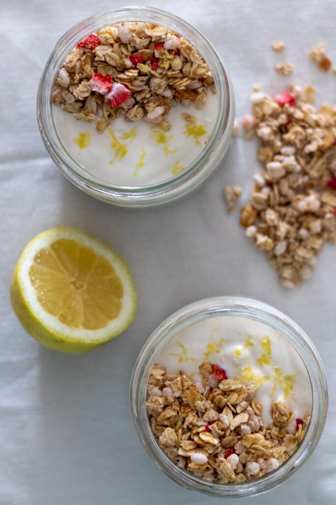 Two small mason jars filled with chia pudding topped with plant-based yogurt, granola and lemon zest with half a lemon and granola on the side.