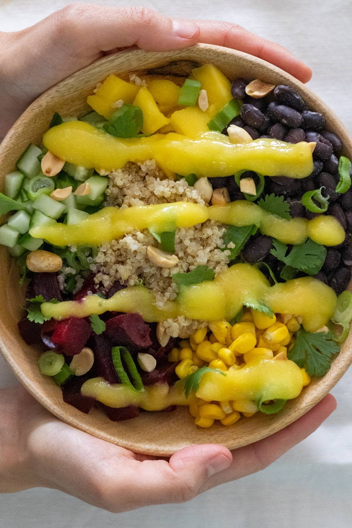 Two hands hold a wooden bowl filled with cooked quinoa, diced cucumber, black beans, corn, cubed beets, cubed mango,  peanuts, spring onion, cilantro and mango sauce.