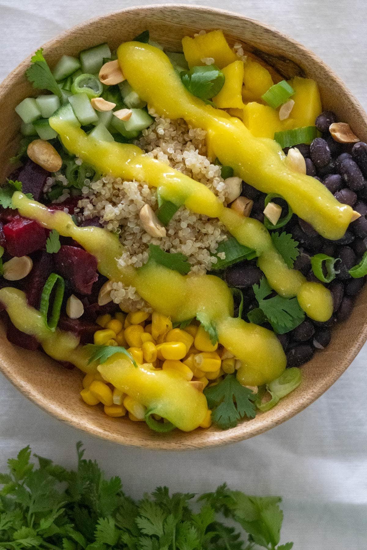 Cooked quinoa, diced cucumber, black beans, corn, cubed beets and cubed mango in a wooden bowl topped with mango sauce, peanuts, spring onion and cilantro.