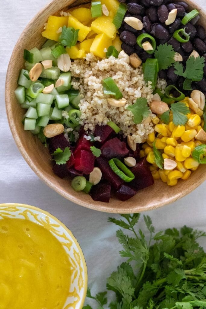 Cooked quinoa, diced cucumber, black beans, corn, cubed beets and cubed mango in a wooden bowl topped with peanuts, spring onion and cilantro. With mango sauce and cilantro on the side.