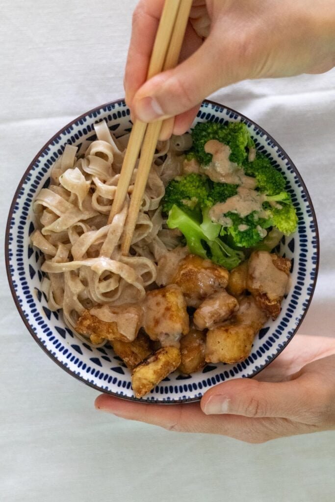 A hand holding chopsticks above a bowl with rice noodles, broccoli and crispy tofu covered in peanut sauce.