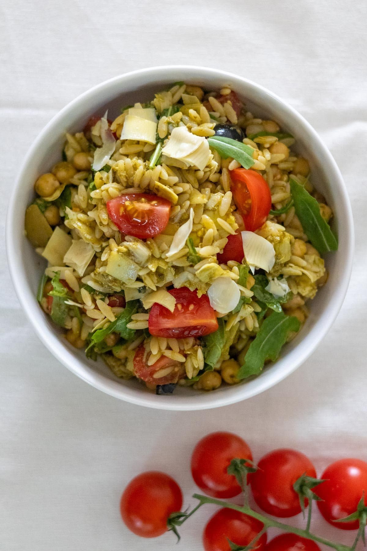 A white bowl filled with cooked orzo, chickpeas, cherry tomatoes, artichoke hearts, arugula, olives and green pesto. 