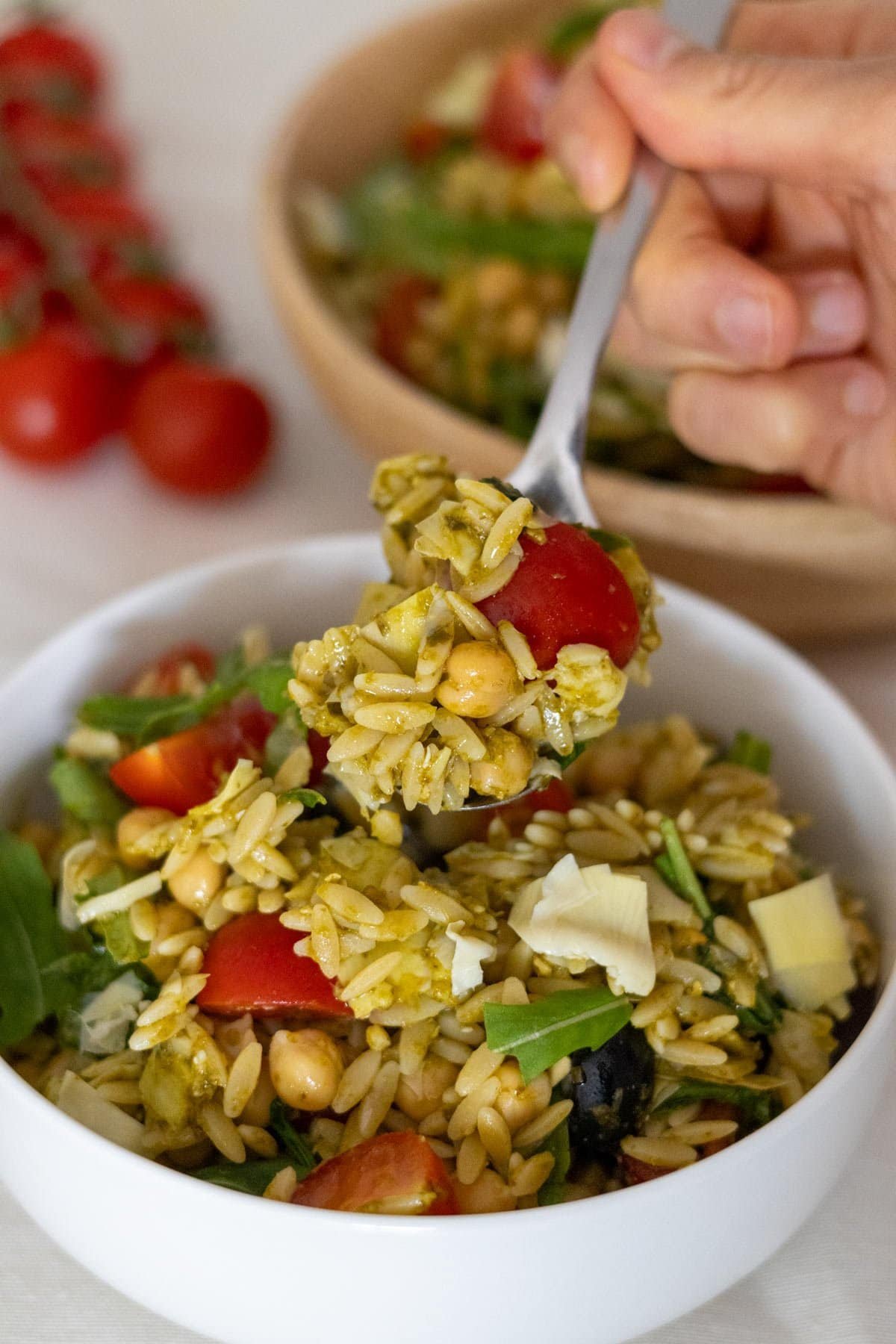 A hand holds a spoon filled with the vegan orzo salad above a bowl with more of the salad.