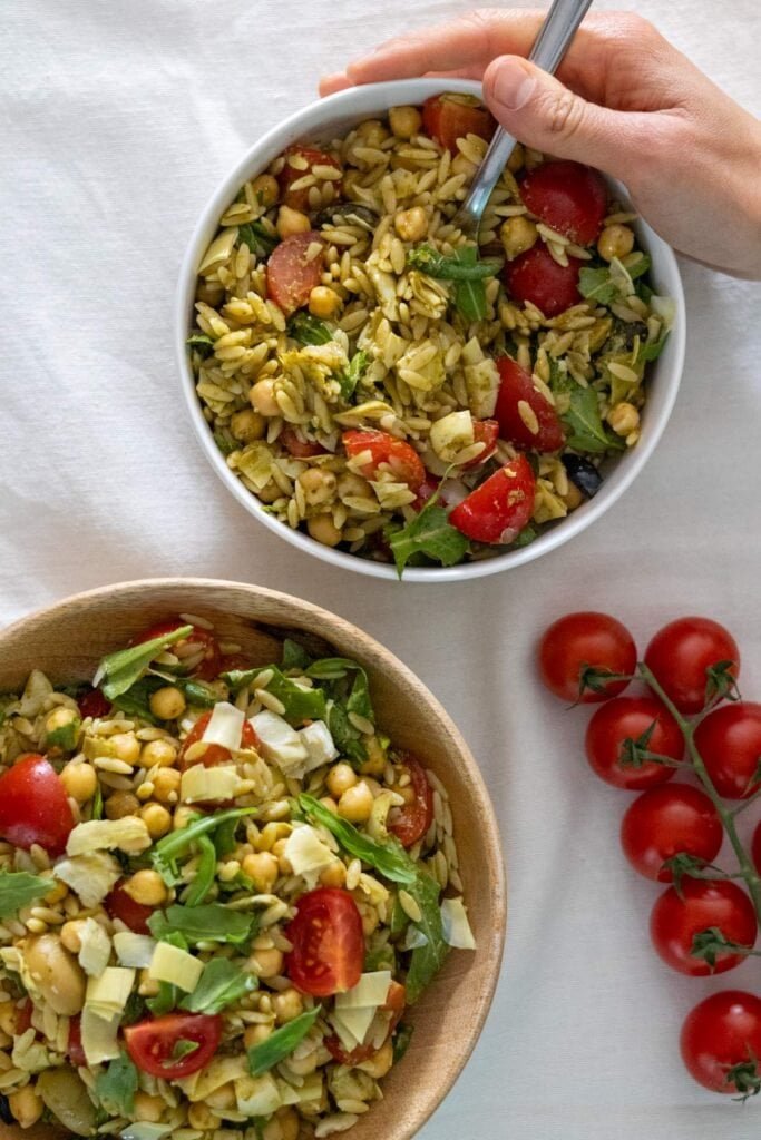 Two bowls filled with orzo, chickpeas, cherry tomatoes, arugula, olives and artichoke hearts. With some cherry tomatoes on the side.