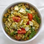 A white bowl filled with cooked orzo, cherry tomatoes, arugula, chickpeas, artichoke hearts, olives and green pesto.