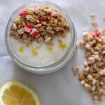 A small mason jar filled with chia pudding topped with plant-based yogurt, granola and lemon zest with half a lemon and granola on the side.