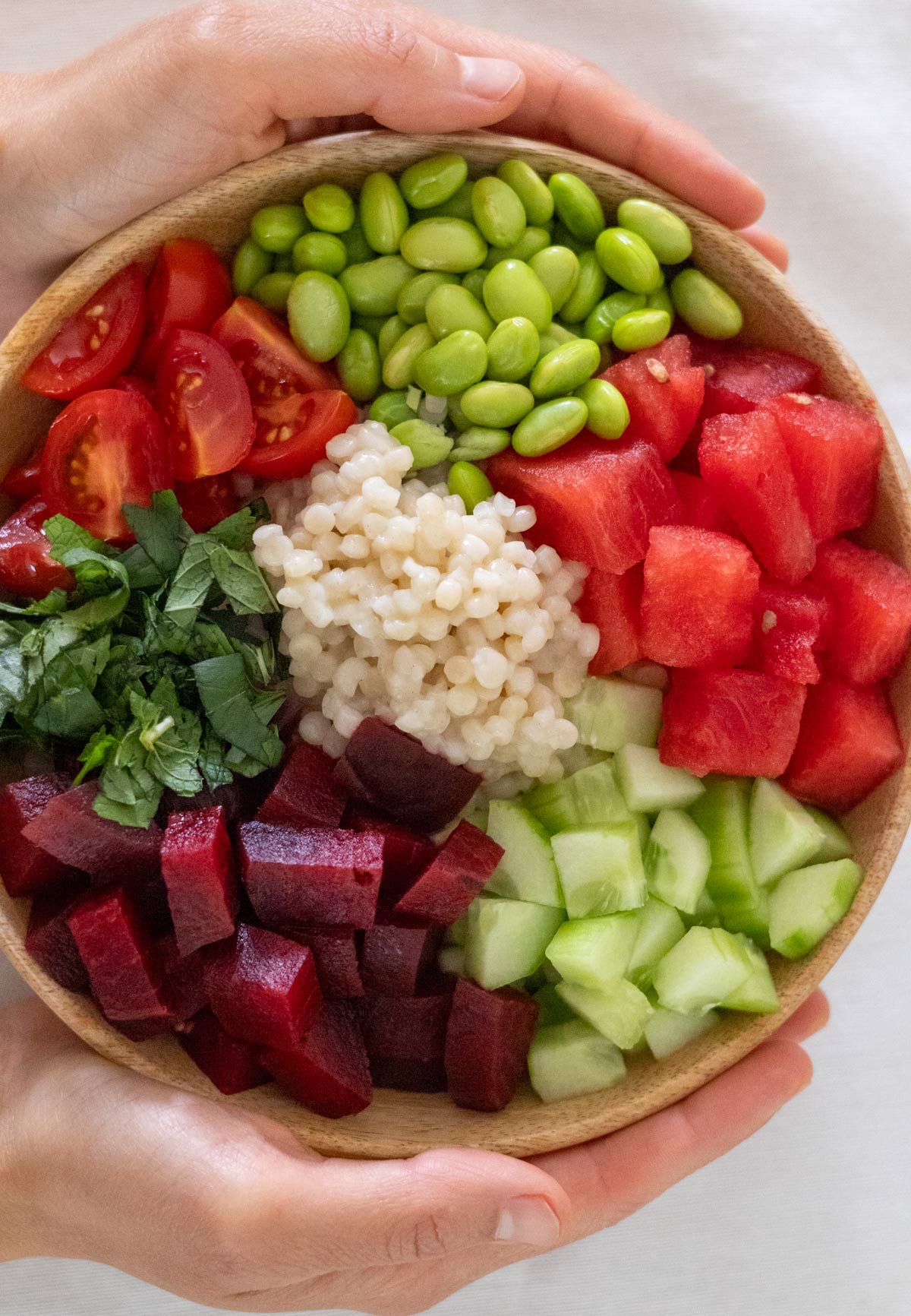 Two hands hold a wooden bowl filled with edamame, cubes of watermelon, cubes beets, pearl couscous, halved cherry tomatoes, cucumber, fresh mint and Thai Basil.