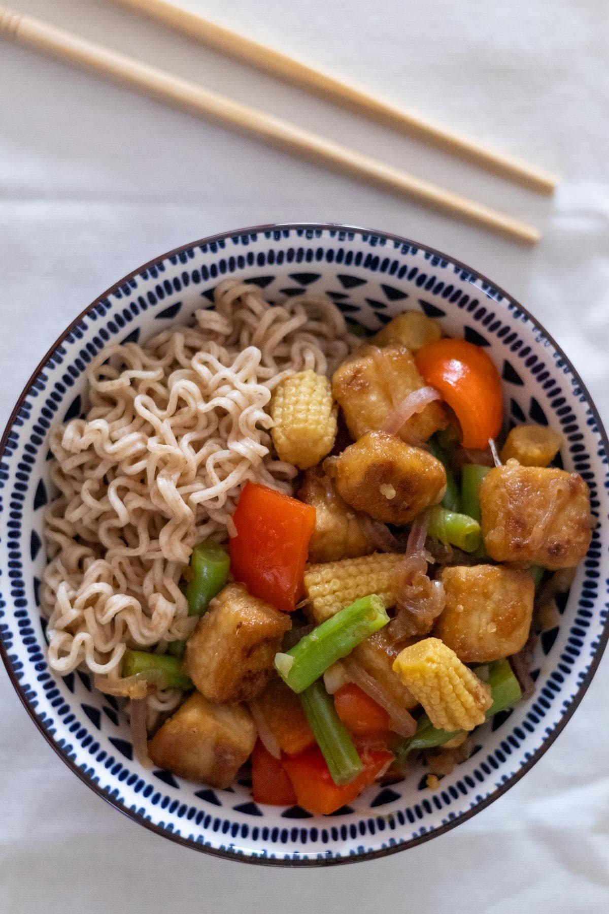 Stir-fried tofu, red bell pepper, baby corn, green beans and red onion served with whole wheat noodles in a blue bowl with chopsticks on the side.