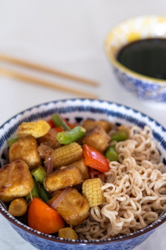 Stir-fried tofu, baby corn, green beans, red onion and red bell pepper with whole wheat noodles in a blue bowl with chopsticks and soy sauce on the side.