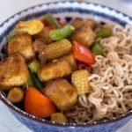 A blue bowl filled with stir-fried tofu, baby corn, red bell pepper, green beans, red onion and cooked whole wheat noodles.